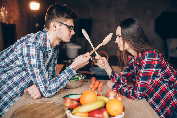Playful young couple fight with wooden spoons on the kitchen while cooking breakfast. Man and woman preparing vegetable salad, happy family together - Photo, Image