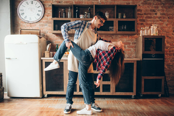 Playful love couple poses on the kitchen. Man in apron and young woman are dancing, funny cooking - Фото, изображение
