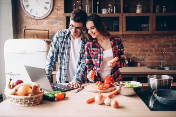 Happy couple looks at a recipe in laptop, vegetable salad cooking on the kitchen. Fresh diet food preparation. Wife and husband prepares romantic dinner together - Foto, Imagem