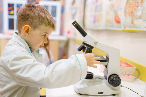 Little boy in uniform sitting at the microscope and playing doctor, playroom. Kid plays medicine worker in imaginary hospital lab, profession learning, childish dream - Valokuva, kuva
