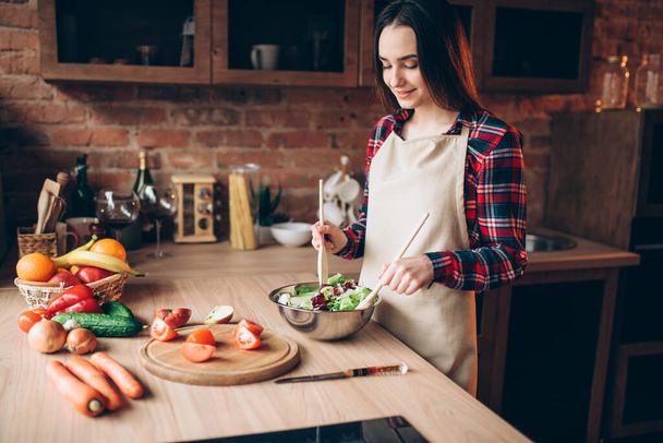 Female cook holds wooden spoon, vegetable salad cooking on the kitchen. Fresh diet food preparation. Wife prepares romantic dinner for her husband - Photo, image