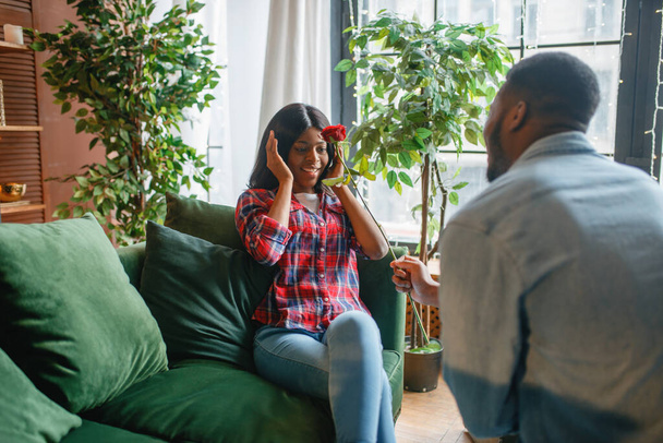 Young black woman with red rose sitting on sofa, romantic meeting at home. Happy african american female person on the couch in living room - Photo, Image