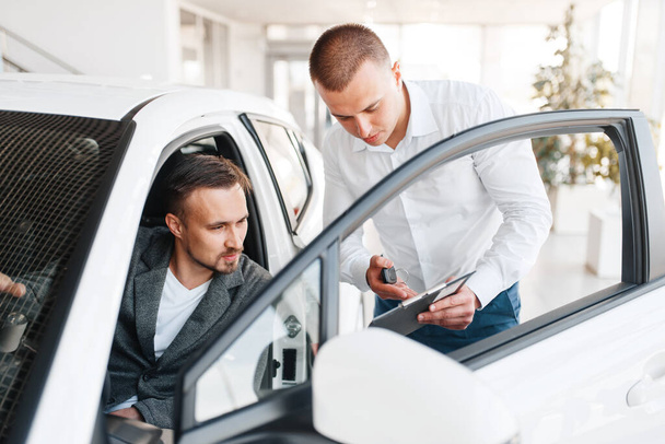 Manager shows to buyer price on new car in showroom. Male customer choosing vehicle in dealership, automobile sale, auto purchase - Fotoğraf, Görsel