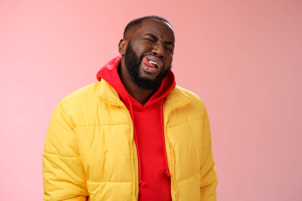 Brutaal stijlvolle grappige Afrikaanse bebaarde man in gele trendy jas rode hoodie show tong brutale flirterige blik knipogen camera proberen indruk te maken vrouw doen alsof macho, staande roze achtergrond - Foto, afbeelding