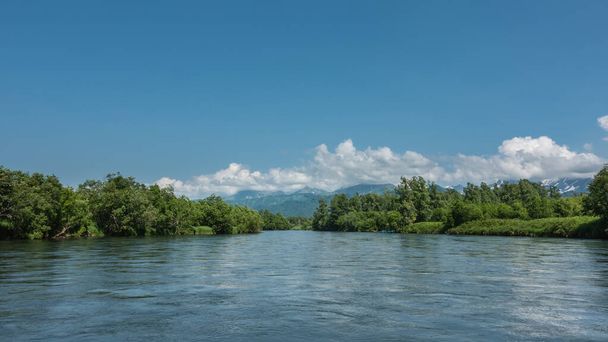 A peaceful blue river flows between the banks with lush green vegetation. In the distance, against the background of the azure sky and clouds, a mountain range is visible. Kamchatka - Photo, Image
