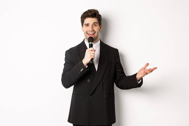 Portrait of handsome man in black suit singing a song, holding microphone and giving speech, standing against white background - Φωτογραφία, εικόνα