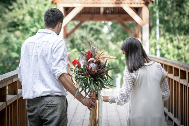 Όμορφο ζευγάρι αγάπης σε έξυπνα ρούχα με ένα μπουκέτο από εξωτικά λουλούδια σε μια ξύλινη γέφυρα, θολή φόντο. - Φωτογραφία, εικόνα