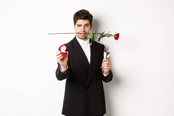 Страстный молодой человек в костюме делает предложение, держит розу в зубах и бокале шампанского, показывает обручальное кольцо, просит выйти за него замуж, стоя на белом фоне - Фото, изображение