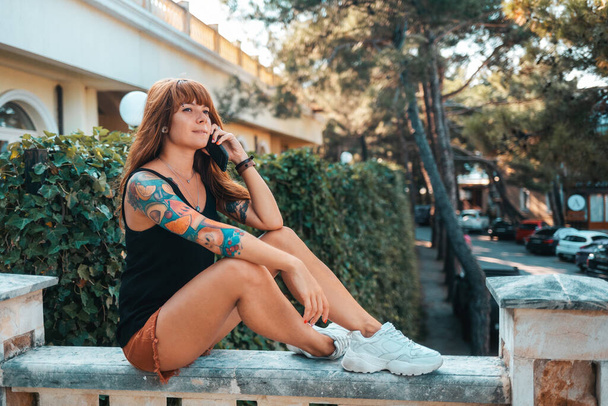 Mujer joven bonita con un tatuaje de manga, relajarse sentado en una escalera de piedra. Mujer hablando en el smartphone. El concepto de comunicación moderna. - Foto, imagen