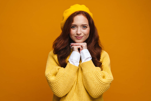Happy young woman with long red hair in sweater and beret standing over yellow wall background - Фото, зображення