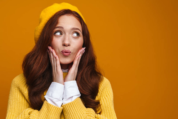 Happy young woman with long red hair in sweater and beret standing over yellow wall background, looking aside - Φωτογραφία, εικόνα