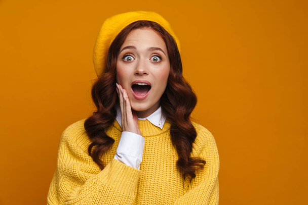 Happy excited young woman with long red hair in sweater and beret standing over yellow wall background, looking at camera - Fotoğraf, Görsel