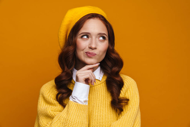 Pensive young woman with long red hair in sweater and beret standing over yellow wall background, looking aside - Фото, зображення