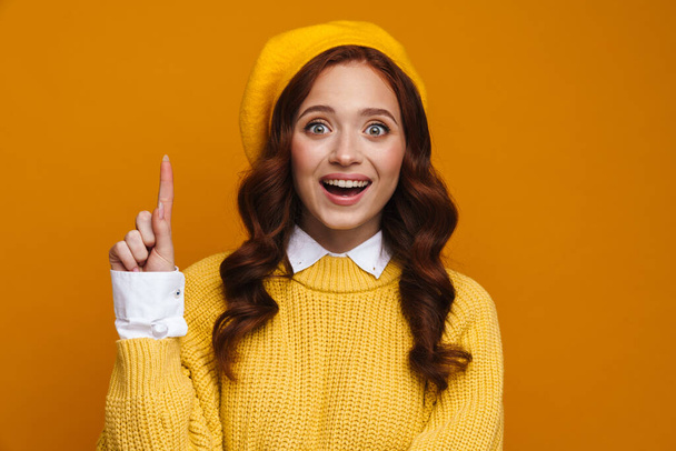 Happy young woman with long red hair in sweater and beret standing over yellow wall background, presenting copy space, pointing aside - Zdjęcie, obraz