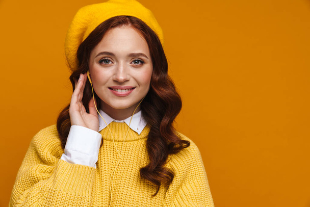 Happy young woman with long red hair in sweater and beret standing over yellow wall background, looking at camera - Zdjęcie, obraz