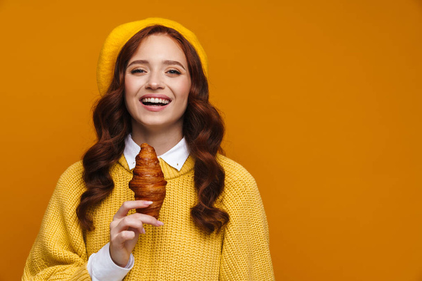 Happy young woman with long red hair in sweater and beret standing over yellow wall background, holding croissant - Foto, Imagen