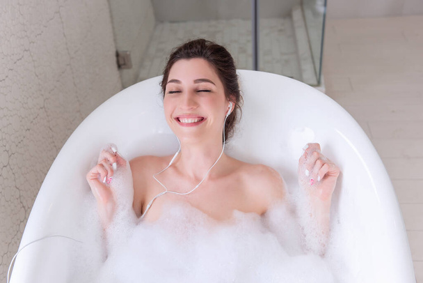 Young millennial woman in headphones listening to music in the bathroom. Happy girl with bun of hair dances in water with white foam, has fun, sings. Spa treatments, relaxing in the morning on weekend - Fotografie, Obrázek