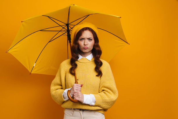Happy young woman with long red hair wearing beret and sweater standing under umbrella over yellow background - Foto, afbeelding