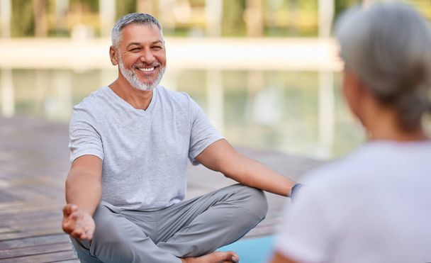 Szczęśliwy pozytywny dojrzały mężczyzna siedzący w pozycji lotosu do jogi z rękami w geście mudry i uśmiechnięty podczas porannej praktyki medytacyjnej z żoną na świeżym powietrzu w parku, para seniorów medytująca na świeżym powietrzu - Zdjęcie, obraz