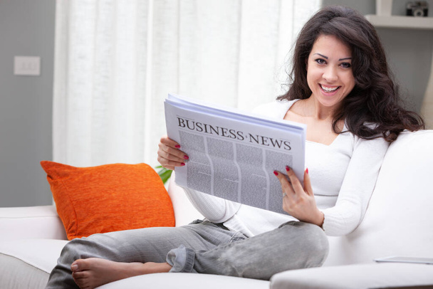Счастливая босиком женщина сидит читает деловую газету на диване дома и смотрит в камеру с сияющей улыбкой - Фото, изображение