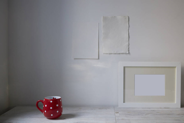 Estilo escandinavo. Design de interiores. Uma xícara vermelha no ponto, uma moldura para uma foto estão na mesa. Duas folhas de papel em branco são anexadas à parede. Espaço vazio para texto - Foto, Imagem