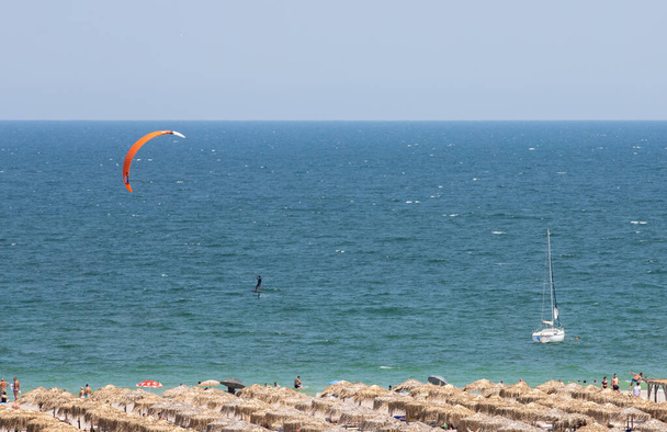 MAMAIA, ROMANIA - Jul 30, 2021: a high angle shot of beach with straw canopies, sailboat, paraglider  on sea in mamaia, romania - Fotoğraf, Görsel