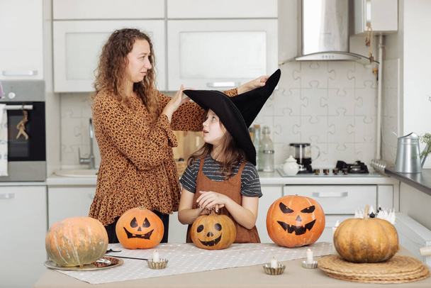 Happy family: mom and daughter celebrate Halloween. Merry child in witch carnival costume in the room. Cheerful kid and parent play with pumpkins and black witch hats at home - Фото, изображение