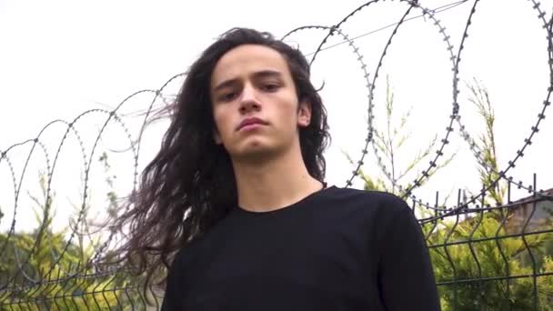 aggressivo guardando lungo dai capelli neri giovane uomo di protesta in piedi accigliato davanti rete metallica con camicia nera - Filmati, video