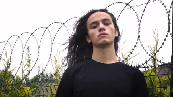 largo negro pelo joven protesta hombre de pie frunciendo el ceño delante de la red de alambre con camisa negra - Imágenes, Vídeo