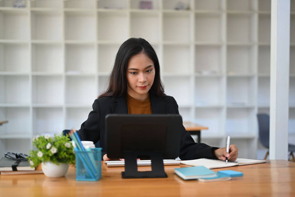 魅力的なビジネス女性は、コンピュータタブレットで作業に集中し、ノートブックにメモを作る. - 写真・画像