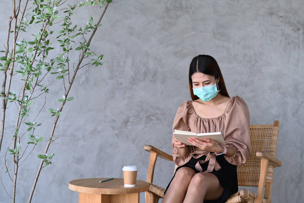 Mladá asijská žena v ochranné masce sedí v kavárně a čtení knihy. - Fotografie, Obrázek