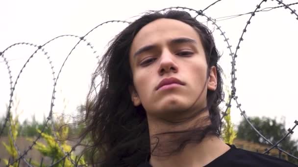 agressif à la recherche de longs cheveux noirs jeune homme de protestation debout scowling en face de treillis métallique - Séquence, vidéo