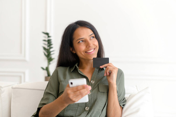 Serene sorrindo sonhador mulher indiana segurando cartão de crédito e smartphone - Foto, Imagem