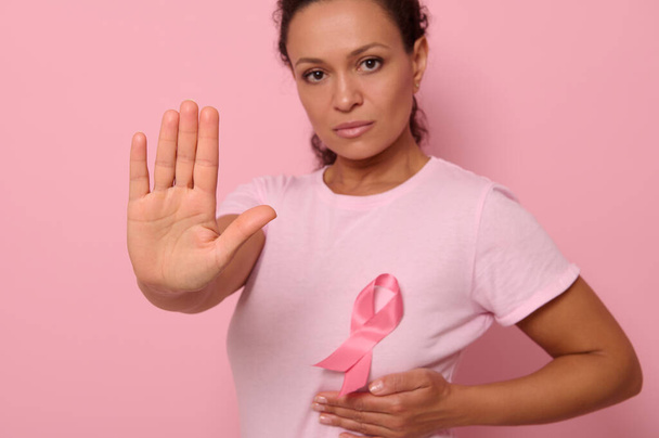 Pembe tişört giyen ve kanser farkındalığı kurdelesi takan bulanık bir kadının kendine güvenen portresi. Kadının el hareketlerine odaklan. Pembe arka planda fotokopi alanı ile izole edilmiş. Kanser Günü - Fotoğraf, Görsel