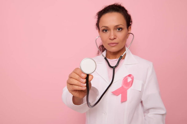 Focus sur le phonendoscope entre les mains d'un beau médecin hispanique métis en manteau médical avec ruban de satin rose, symbole de la Journée mondiale de sensibilisation au cancer du sein, Octobre rose. Concept de santé de la femme - Photo, image