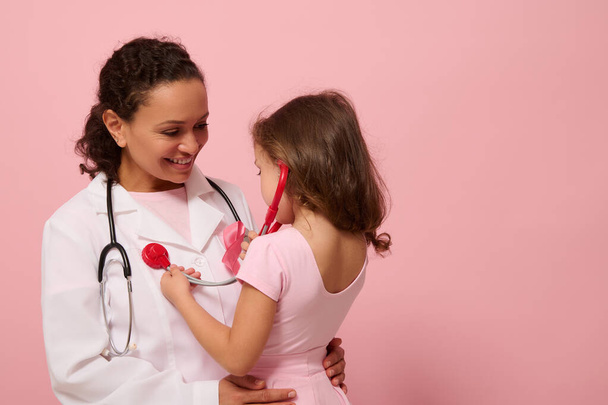 Kleines Mädchen in pinkfarbener Kleidung benutzt Stethoskop, hört Brust und Lungen atmen des Arztes mit rosa Schleife auf der Brust. Konzept eines Bildungsprogramms zur Unterstützung von Brustkrebspatientinnen. Kopierraum - Foto, Bild