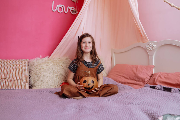 Söpö pieni tyttö leikkii Halloween kurpitsa sängyllä hänen vaaleanpunainen huone. Tyttö katsoo kameraan ja hymyilee. halloween puolue kotona. - Valokuva, kuva
