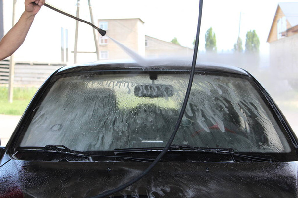 ένας άνθρωπος πλένει ανεξάρτητα ένα αυτοκίνητο kercher / φωτογραφία ένας άνθρωπος πλένει ανεξάρτητα ένα αυτοκίνητο. Έχει ένα κέρατο στα χέρια του. ένα πίδακα νερού υψηλής πίεσης. Το αυτοκίνητο είναι μαύρο.. - Φωτογραφία, εικόνα