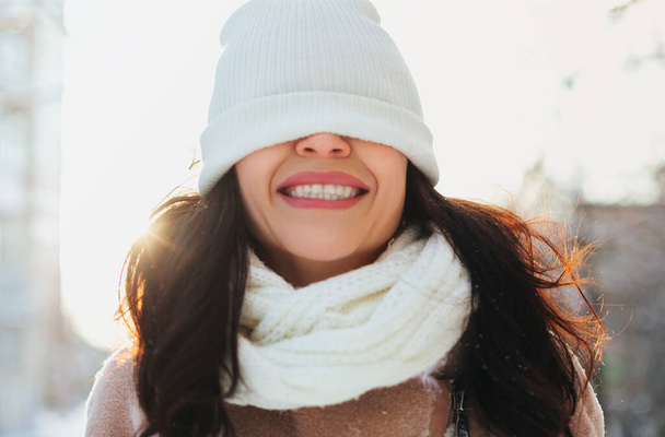 Optimista fiatal nő meleg kalappal a szemén mosolyog, miközben szórakozik az utcán napsütéses téli napon - Fotó, kép