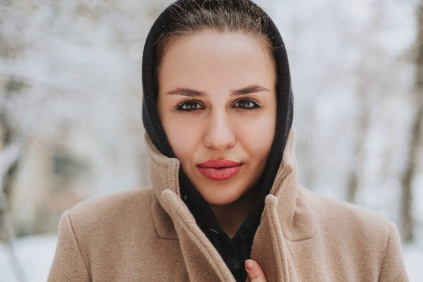 Молодая позитивная женщина со смешной улыбкой на лице стоит на улице в бежевом пальто и черном шарфе на голове, дрожа от зимней холодной погоды на фоне деревьев, покрытых толстым слоем снега - Фото, изображение