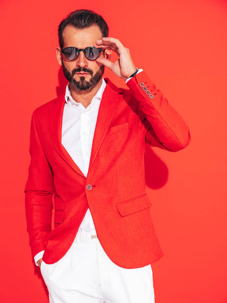 Portrait of handsome confident stylish hipster lambersexual model. Sexy modern man dressed in elegant suit. Fashion male posing in studio near red wall in sunglasses - Fotoğraf, Görsel