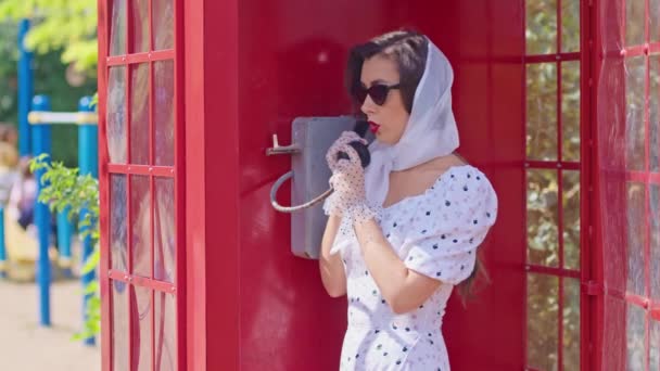 belle jeune femme parle joyeusement au téléphone dans une cabine téléphonique rouge de style anglais. fille habillée dans une robe blanche et des lunettes. - Séquence, vidéo