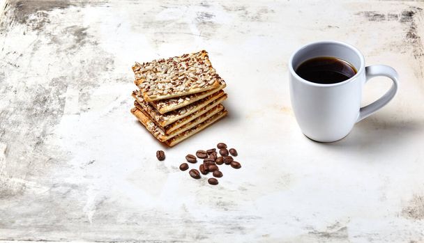 White cup with coffee, coffee beans and crunchy crispbread on a wooden background, closeup. Tasty breakfast, morning routine concept - Photo, image