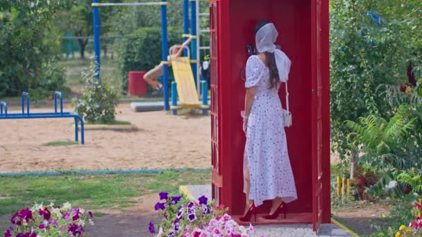όμορφη νεαρή γυναίκα μιλάει ευτυχισμένα στο τηλέφωνο σε ένα αγγλικό στυλ κόκκινο τηλεφωνικό θάλαμο. κορίτσι ντυμένη με λευκό φόρεμα και γυαλιά. - Πλάνα, βίντεο