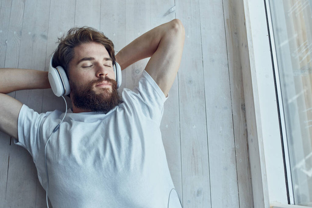 homem de t-shirt branca deitado perto da janela usando tecnologia de fones de ouvido - Foto, Imagem