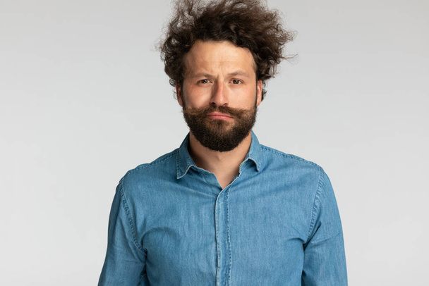 portrait of handsome man in denim shirt with curly hair posing and making faces on grey background in studio - Photo, Image