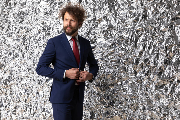 sexy barbudo hombre de negocios en traje azul desabotonando traje y mirando a un lado en el fondo de papel de aluminio en el estudio - Foto, imagen
