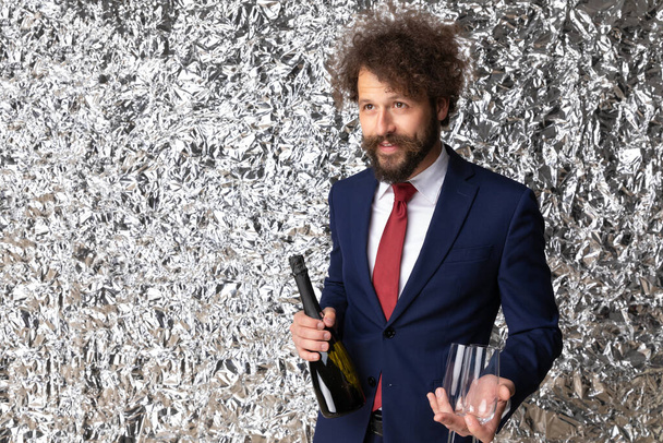 portrait of happy young man in navy blue suit holding champagne bottle and glasses, looking up and side and posing in front of tinfoil background in studio - Photo, Image