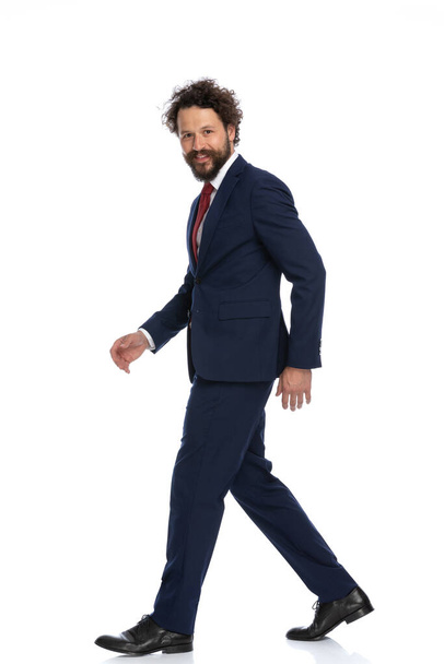 boční pohled na atraktivního podnikatele kráčejícího, dívajícího se do kamery a usmívajícího se na bílém pozadí - Fotografie, Obrázek