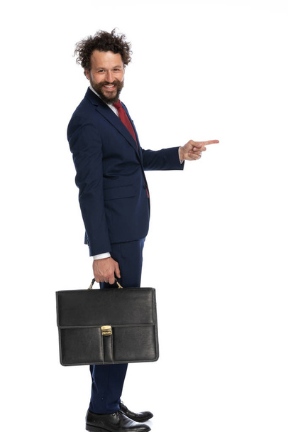 zijaanzicht van een blije zakenman die naar de zijkant wijst, glimlacht en een aktetas op witte achtergrond vasthoudt - Foto, afbeelding